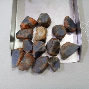 May include: A collection of rough, natural blue sapphires with some orange inclusions. The sapphires are various sizes and shapes, and are displayed on a silver tray.