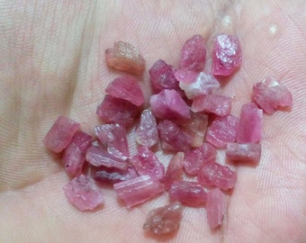 May include: A collection of pink ruby rough crystals. The crystals are various sizes and shapes, with some being long and thin, while others are more rounded. The crystals are all a vibrant pink color.