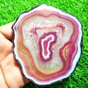 May include: A polished agate slice featuring concentric bands of deep magenta, soft pink, and translucent white. The center displays a crystalline geode formation with jagged edges, surrounded by smooth, layered rings of varying color intensity.