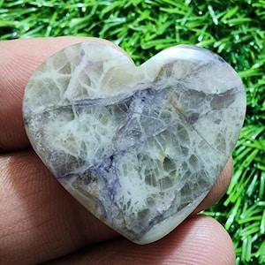 May include: A heart-shaped stone with a marbled pattern of white, gray, and purple. The stone has a polished surface and is held in a hand against a background of green grass. The stone's unique veining creates a natural, artistic design.