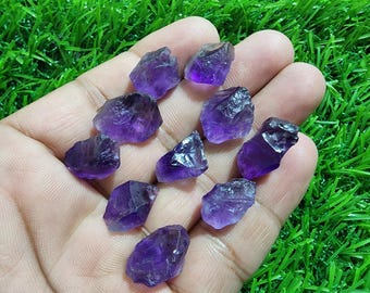Peut inclure: Plusieurs cristaux d'améthyste bruts de différentes formes et tailles, présentés dans une main sur un fond d'herbe verte. Les cristaux affichent une teinte violette foncée, avec des zones plus sombres.