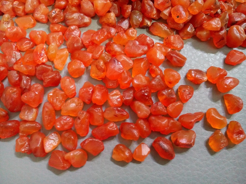K&ouml;nnte beinhalten: Eine Sammlung kleiner, glatter, orangefarbener Karneol-Steine. Die Steine sind unregelm&auml;&szlig;ig geformt und haben eine nat&uuml;rliche, polierte Oberfl&auml;che.