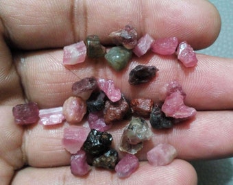 May include: A collection of small, rough-cut gemstones in various shades of pink, red, and brown. The gemstones are held in a hand, showcasing their natural, irregular shapes and subtle variations in color and texture. These raw gemstones are ideal for jewelry making.