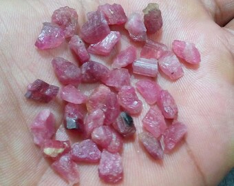 May include: A handful of rough-cut, pink tourmaline gemstones. The stones vary in size and shape, displaying a range of pink hues, from light to deep rose. The gemstones are held in an open hand, showcasing their natural, unpolished state.