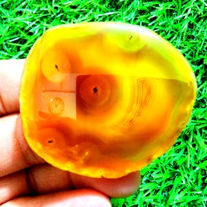 May include: A polished agate stone, displaying a mix of yellow, orange, and brown tones. The stone has a rounded shape and features concentric banding patterns. The surface reflects light, showcasing the natural textures and variations of the gemstone. The stone is translucent.