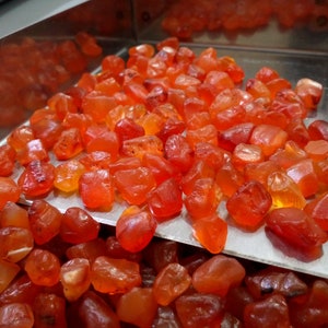 K&ouml;nnte beinhalten: Nahaufnahme eines Metalltabletts gef&uuml;llt mit kleinen, rauen, orange-roten Karneol-Edelsteinen. Das Tablett liegt auf einem Haufen &auml;hnlicher Karneol-Steine.