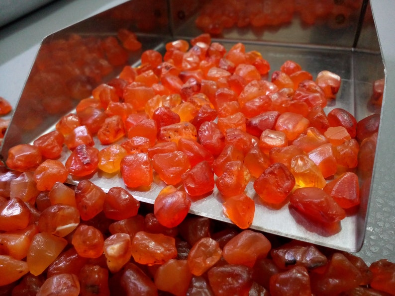 K&ouml;nnte beinhalten: Nahaufnahme eines Metalltabletts gef&uuml;llt mit kleinen, rauen, orange-roten Karneol-Edelsteinen. Das Tablett liegt auf einem Haufen &auml;hnlicher Karneol-Steine.