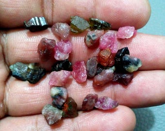 May include: A collection of raw gemstones in various colors, including pink, red, black, and green. The gemstones are small, irregular shapes, and appear to be uncut. The image shows the gemstones resting in a person's hand.