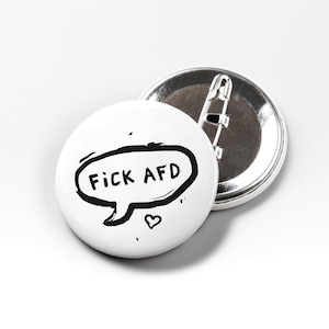 Könnte beinhalten: Zwei weiße runde Ansteckbuttons. Auf einem Button steht der Text "FICK AFD" in einer schwarzen Sprechblase mit einem kleinen Herz. Der andere Button ist silberfarben und hat eine Sicherheitsnadel auf der Rückseite.