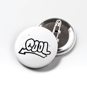 Könnte beinhalten: Zwei weiße, runde Ansteckbuttons. Einer zeigt das Wort "COOL" in schwarzer Graffiti-Schrift mit einem nach links zeigenden Pfeil. Der andere ist silberfarben mit Sicherheitsnadelverschluss. Die Buttons befinden sich auf weißem Hintergrund.