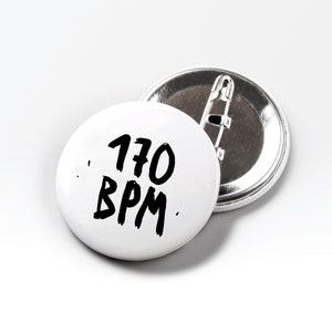 Może przedstawiać: Dwa białe, okrągłe buttony. Przedni button ma czarny napis "170 BPM" w stylu ręcznym. Widoczny jest tył jednego buttonu, pokazujący srebrną agrafkę. Buttony znajdują się na białym tle.