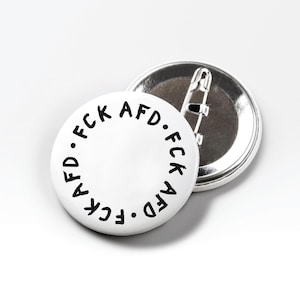 May include: Two white round buttons with black text. The text on the front of the button reads "FCK AFD" in a circular pattern. The back of one button is silver with a safety pin clasp.