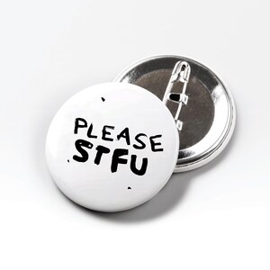 May include: Two white round badges with metal backs. One badge is face up, displaying the text "PLEASE STFU" in black, handwritten-style lettering. The other badge is face down, showing the metal pin backing.