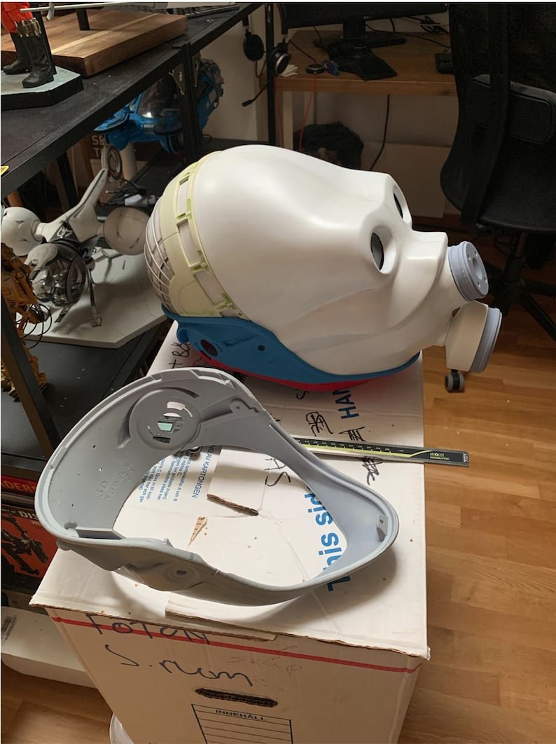 May include: A white and blue helmet with a gray neck piece. The helmet has a large, round, white head piece with two black eye holes and a gray, cylindrical nose piece. The neck piece is a gray, curved piece that fits around the neck.