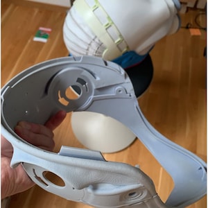 May include: A light gray plastic helmet with a textured surface and several holes. The helmet is designed to fit around the head and neck.
