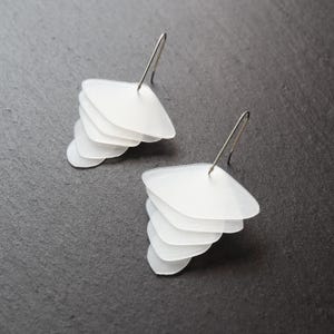 May include: A pair of white, layered earrings with a cascading petal design. Each earring features multiple translucent, petal-shaped elements suspended from a silver-colored hook. The earrings are displayed against a dark gray background.