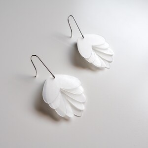 May include: A pair of white, layered petal-shaped earrings with silver-tone hooks. The earrings are made of multiple translucent, oval-shaped pieces, creating a layered effect. The design is minimalist and modern, with a focus on shape and texture.