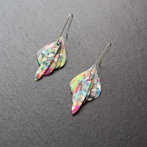 May include: A pair of abstract dangle earrings. Each earring features a layered, leaf-shaped design with a colorful, speckled pattern in shades of pink, green, blue, and yellow. The earrings have silver-colored ear wires.