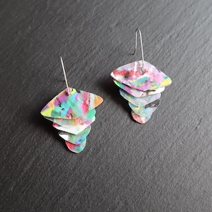 May include: A pair of abstract earrings with a layered design. Each earring features multiple diamond-shaped pieces in a variety of colors, including pink, green, blue, and yellow. The earrings have silver-colored ear wires.