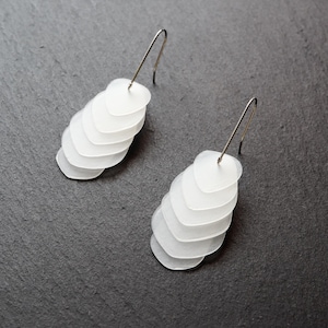 May include: A pair of white, layered earrings with a cascading design. Each earring features multiple heart-shaped pieces that gradually increase in size. The earrings are suspended from silver-colored ear wires. The jewelry is displayed on a dark gray surface.