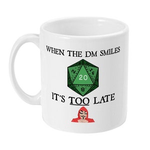 Op de afbeelding: Een witte keramische mok met een groene 20-zijdige dobbelsteen en de tekst "When the DM smiles it's too late". Een rode gekapte figuur met een zwart masker bevindt zich aan de onderkant van de mok.