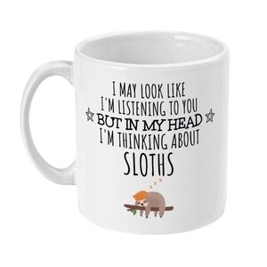 May include: White ceramic mug with a black and white graphic of a sloth sleeping on a branch. The text on the mug reads "I may look like I'm listening to you but in my head I'm thinking about sloths."