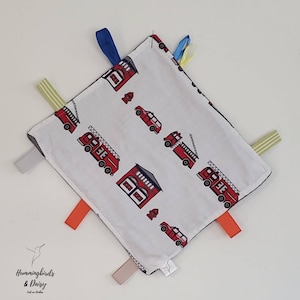 May include: A square baby comforter with a white background patterned with red fire engines and fire stations. The blanket has a black border and colourful ribbons attached to each corner. The Hummingbirds & Daisy logo is in the bottom left corner.