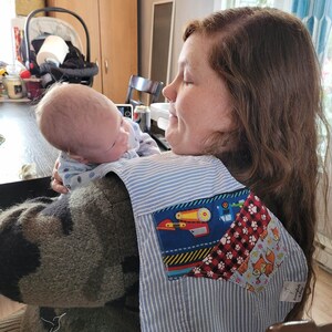 May include: A woman wearing a blue and white striped shirt with a patchwork pocket. The pocket is made of various fabrics with different patterns, including a red and black check, a blue fabric with a red truck, and a pink fabric with a fox. The woman is holding a baby in her arms. The baby is wearing a white and blue striped shirt. The text "Hummingbirds & Daisy" is visible on the shirt.