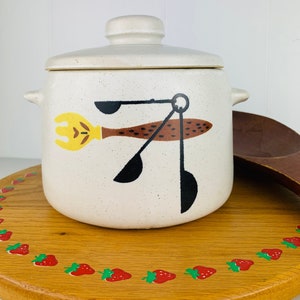May include: White ceramic crock pot with a lid and a brown, yellow, and black design of kitchen utensils. The crock pot is sitting on a wooden surface with a strawberry border.