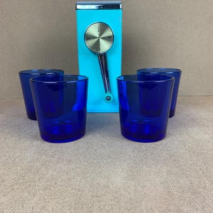 May include: A vintage turquoise ice crusher with a silver handle and dial, accompanied by four cobalt blue drinking glasses. The ice crusher has a chrome top and is set against a neutral background.