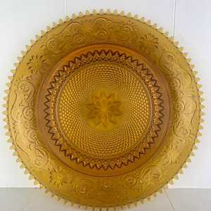 May include: Amber-colored glass plate with intricate embossed floral and starburst patterns. The plate features a scalloped edge and a central star design. A decorative piece for serving or display.