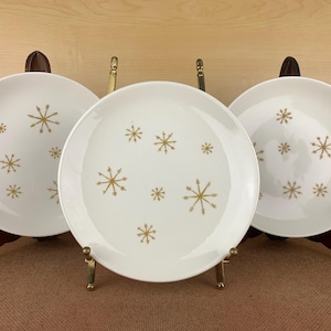 May include: Three white ceramic plates with a gold snowflake design. The plates are displayed on wooden and brass plate stands. The plates have a mid-century modern aesthetic and are suitable for serving food.