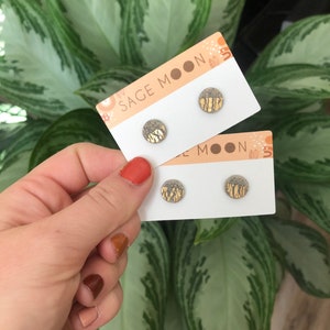 May include: Two pairs of circular stud earrings displayed on white Sage Moon branded cards. Each earring features a textured grey speckled upper half and a metallic gold leaf lower half with vertical brushstroke patterns on the surface.
