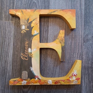 May include: A wooden letter E painted with a whimsical forest scene. The letter is painted in shades of yellow, orange, and brown. The letter is decorated with small white fairy figures, a brown door, and red mushrooms. The letter is personalised with the name "Eleanor".