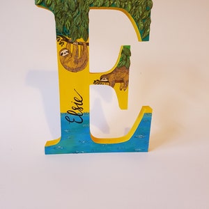 May include: A yellow wooden letter E with a green leafy canopy, two sloths, and the name "Elsie" written in black cursive. The letter is painted with blue water at the bottom.