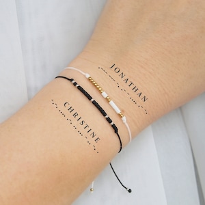 May include: Two friendship bracelets with black and white beads and gold accents. The bracelets are tied around a person's wrist. The words "JONATHAN" and "CHRISTINE" are written in Morse code on the person's arm.