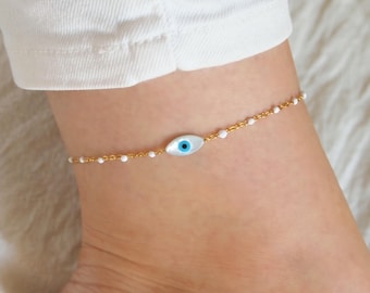 May include: Gold anklet with a white evil eye charm. The anklet is made of small white beads and a delicate gold chain.