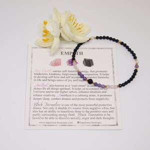 May include: Black, pink, and purple gemstone bracelet with gold accents. The bracelet is on a white card with the text "EMPATH" and descriptions of the stones: Rose Quartz, Amethyst, and Black Tourmaline. The card also includes the website address "www.lapaloma-boutique.com" and the logo "lapaloma.boutique".