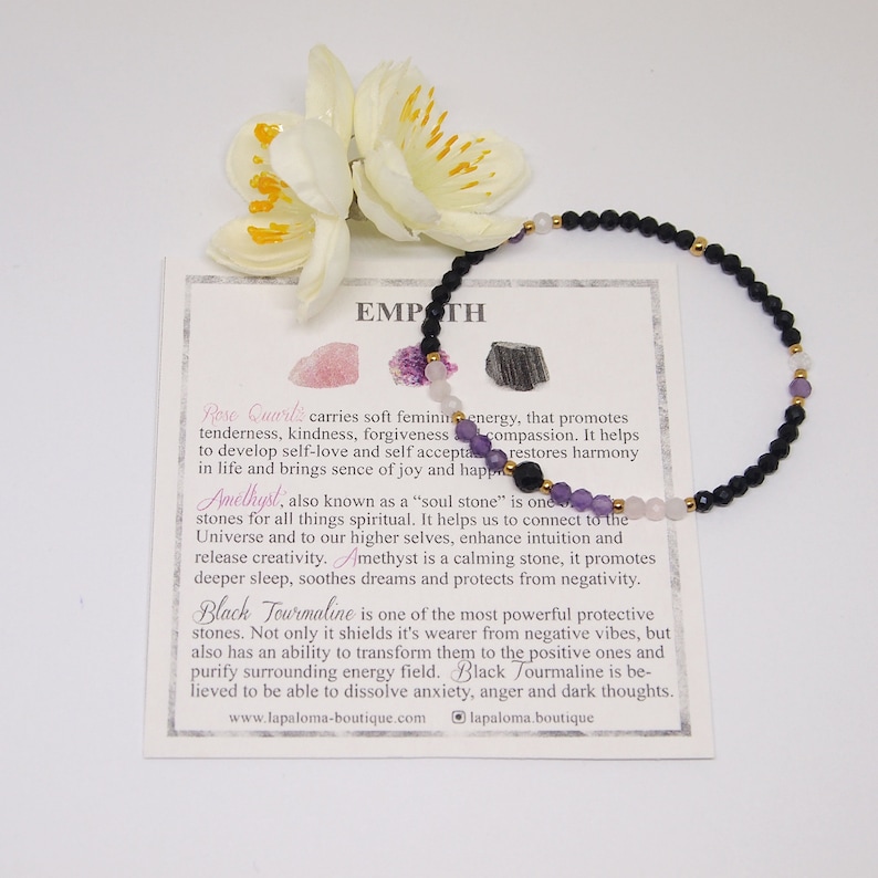 May include: Black, pink, and purple gemstone bracelet with gold accents. The bracelet is on a white card with the text "EMPATH" and descriptions of the stones: Rose Quartz, Amethyst, and Black Tourmaline. The card also includes the website address "www.lapaloma-boutique.com" and the logo "lapaloma.boutique".