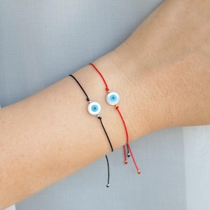 May include: Two evil eye bracelets on a wrist. One bracelet is red string with a gold bead and a blue evil eye charm. The other bracelet is black string with a silver bead and a blue evil eye charm.