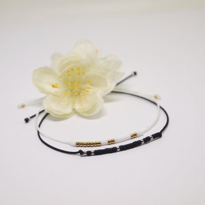 May include: Two friendship bracelets with black and white string and gold beads. The bracelets are tied together with a white flower.