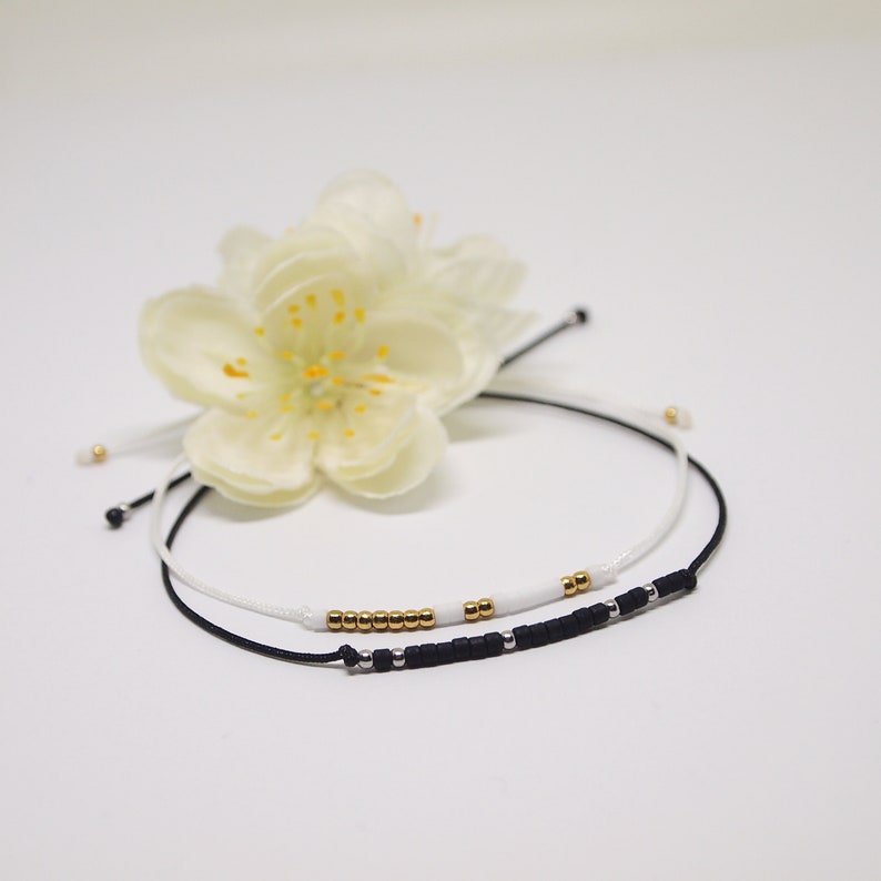 May include: Two friendship bracelets with black and white string and gold beads. The bracelets are tied together with a white flower.