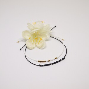 May include: Two beaded bracelets, one black with silver beads and one white with gold beads, on a white background with a white flower.