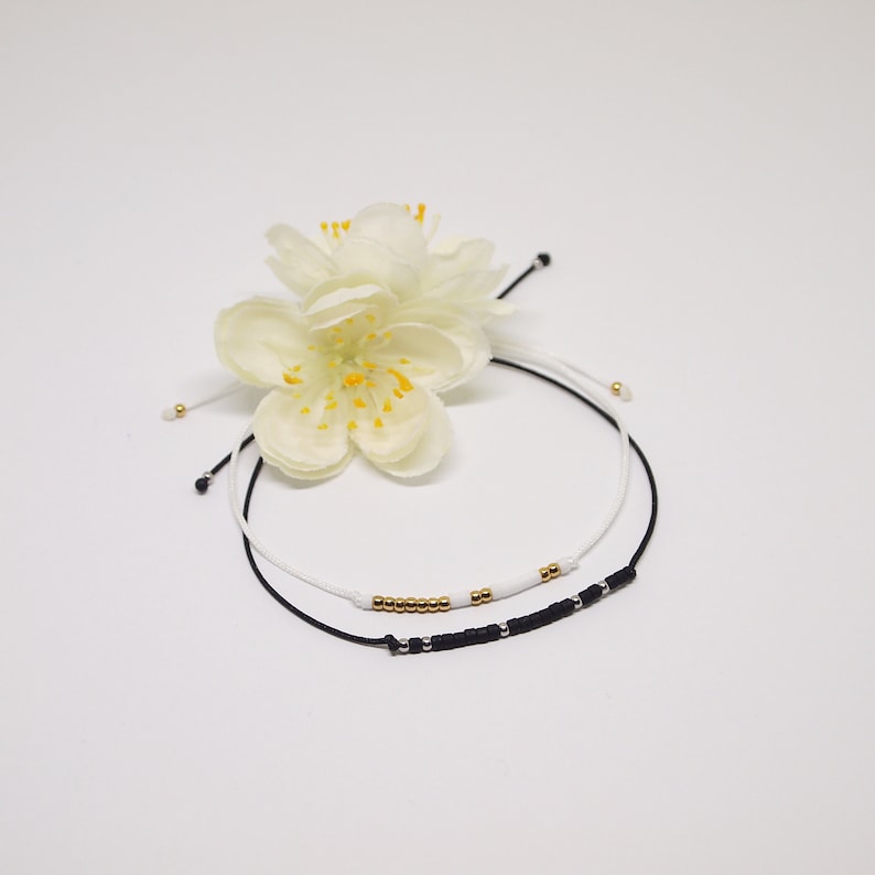 May include: Two beaded bracelets, one black with silver beads and one white with gold beads, on a white background with a white flower.