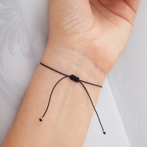 May include: A black adjustable bracelet with silver beads at the ends. The bracelet is tied around a person's wrist.