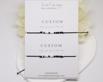 May include: Two black string bracelets with white and gold beads spelling out the word "CUSTOM" in Morse code. The bracelets are displayed on a white card with the text "Secret message morse code bracelet" and "HANDMADE www.lapalomalaboutiqe.com".