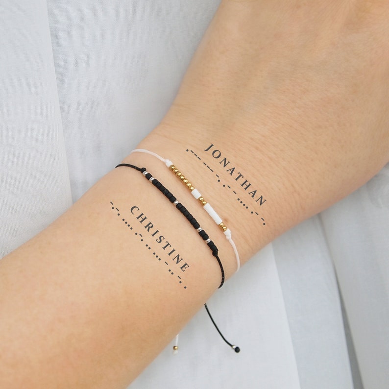 May include: Two friendship bracelets with black and white beads and gold accents. The bracelets are tied around a person's wrist. The words "JONATHAN" and "CHRISTINE" are written in Morse code on the person's arm.