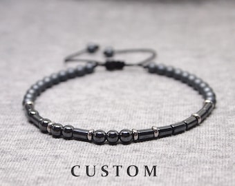 May include: A black beaded bracelet with silver accents and a black cord closure. The bracelet is on a gray fabric background.