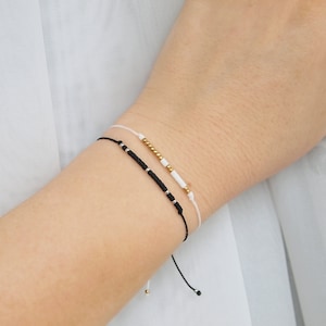 May include: Two adjustable bracelets with black and white beads on a white string. The bracelet on top has gold beads.