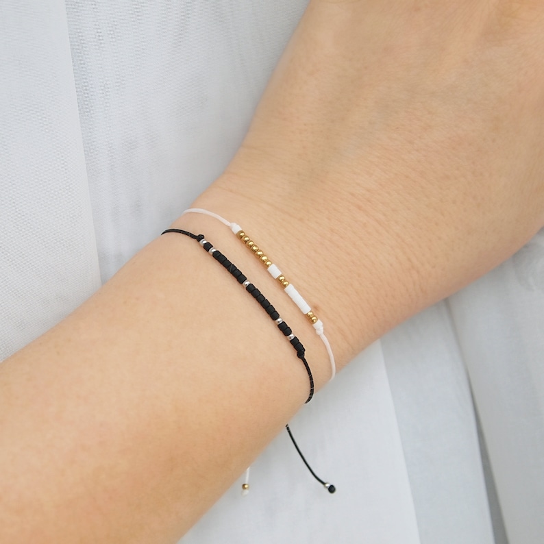 May include: Two adjustable bracelets with black and white beads on a white string. The bracelet on top has gold beads.